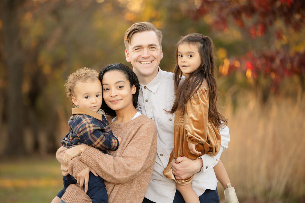 Family looking at the camera smiling with kids being held by parents with fall colors in the background what to wear to a fall mini session
