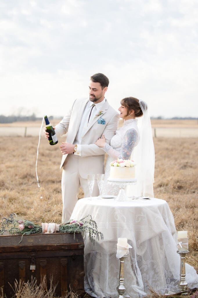 Bride and groom looking at overflowing champagne bottle, near a table with a lace cake at Cedar Lawn Farms in Topeka, Kansas.